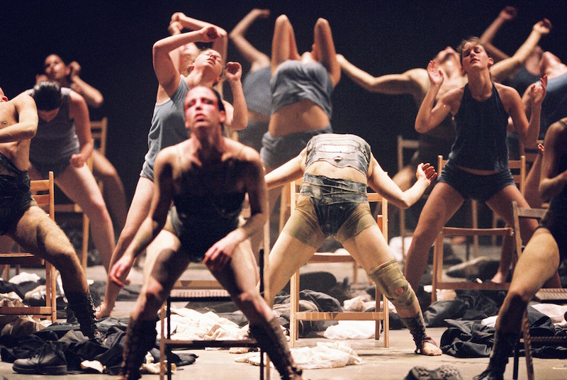 A group of dancers hover over chairs. Shoes and clothers are strewn about. One dancer assumes a dramatic back end with her legs wide apart.